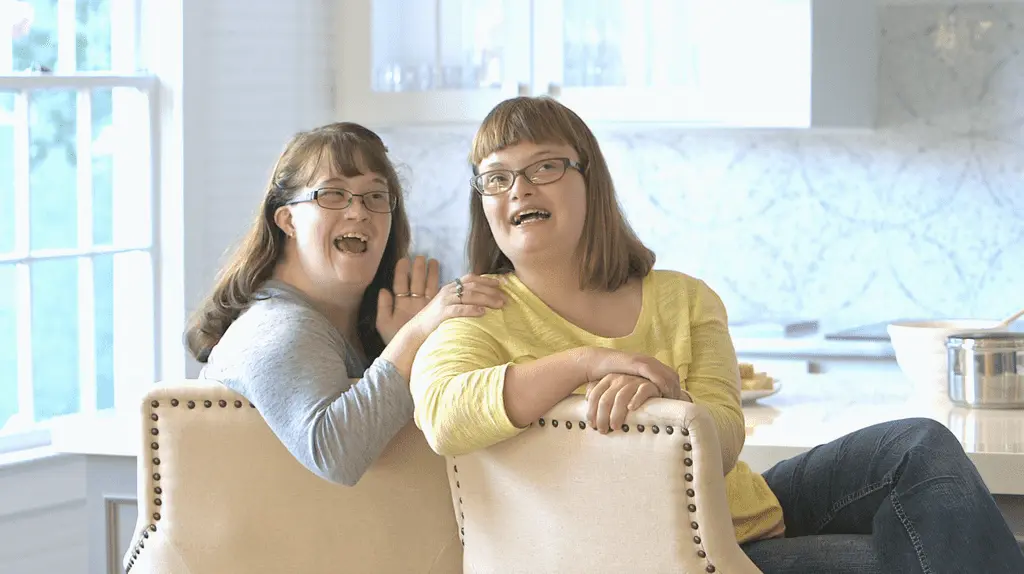 Sisters smiling while sitting on chairs next to one another at the kitchen counter.
